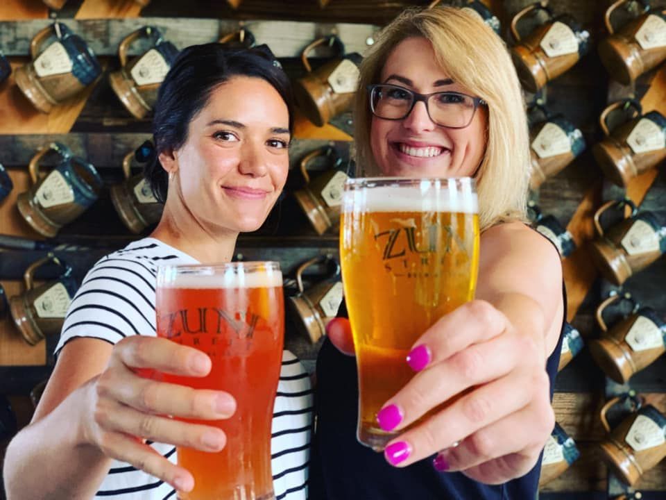 Two women are holding up glasses of beer in front of a wall of mugs.