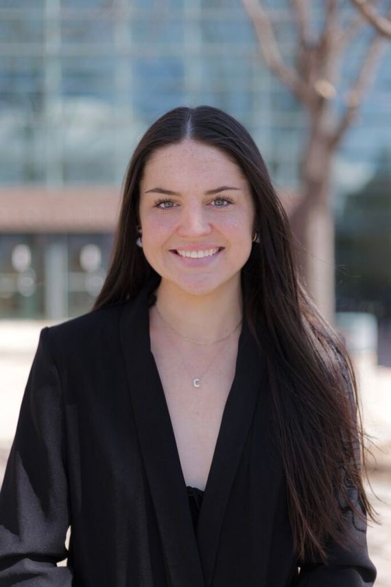 Woman smiling outdoors in front of a building, wearing a black top and long dark hair.