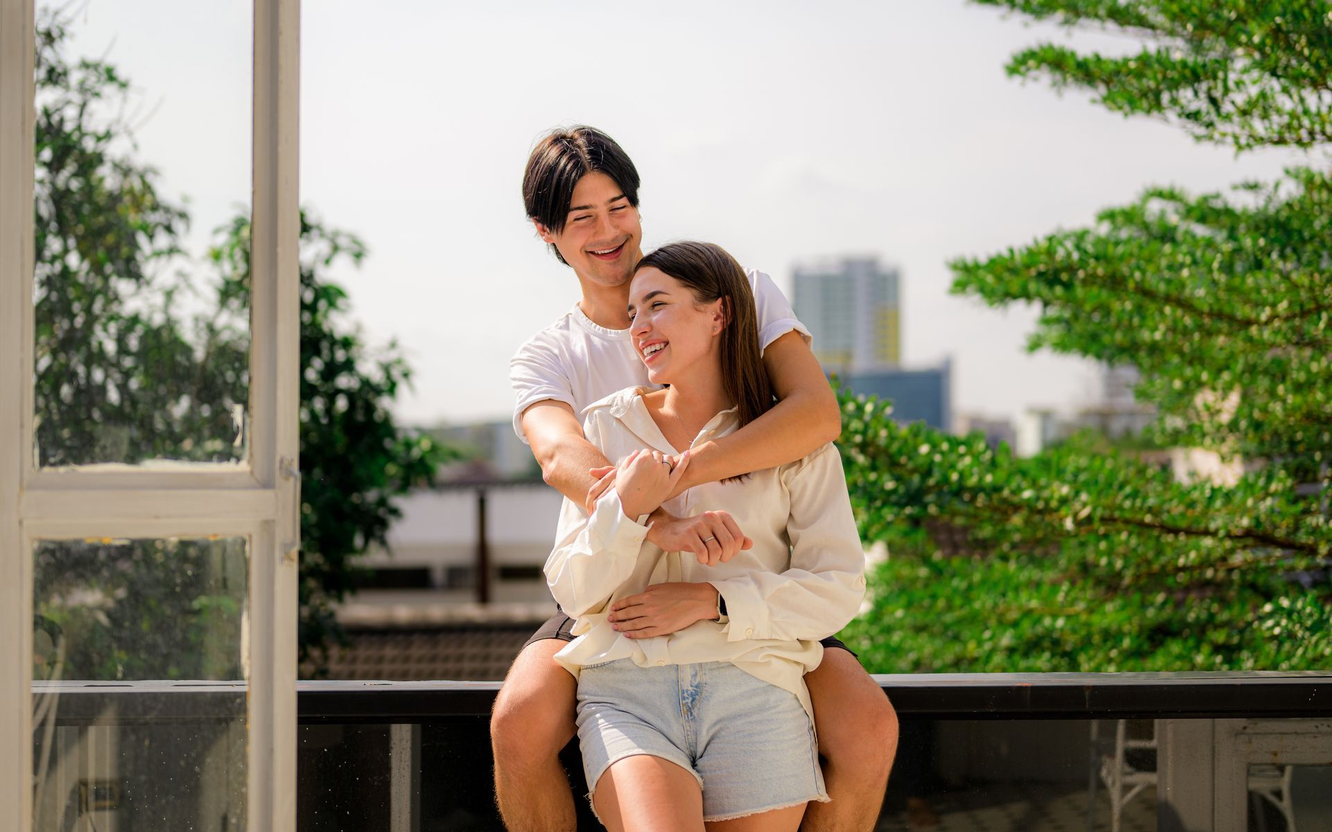 Two people sitting together on a balcony, smiling and hugging, with greenery and city buildings behind them.