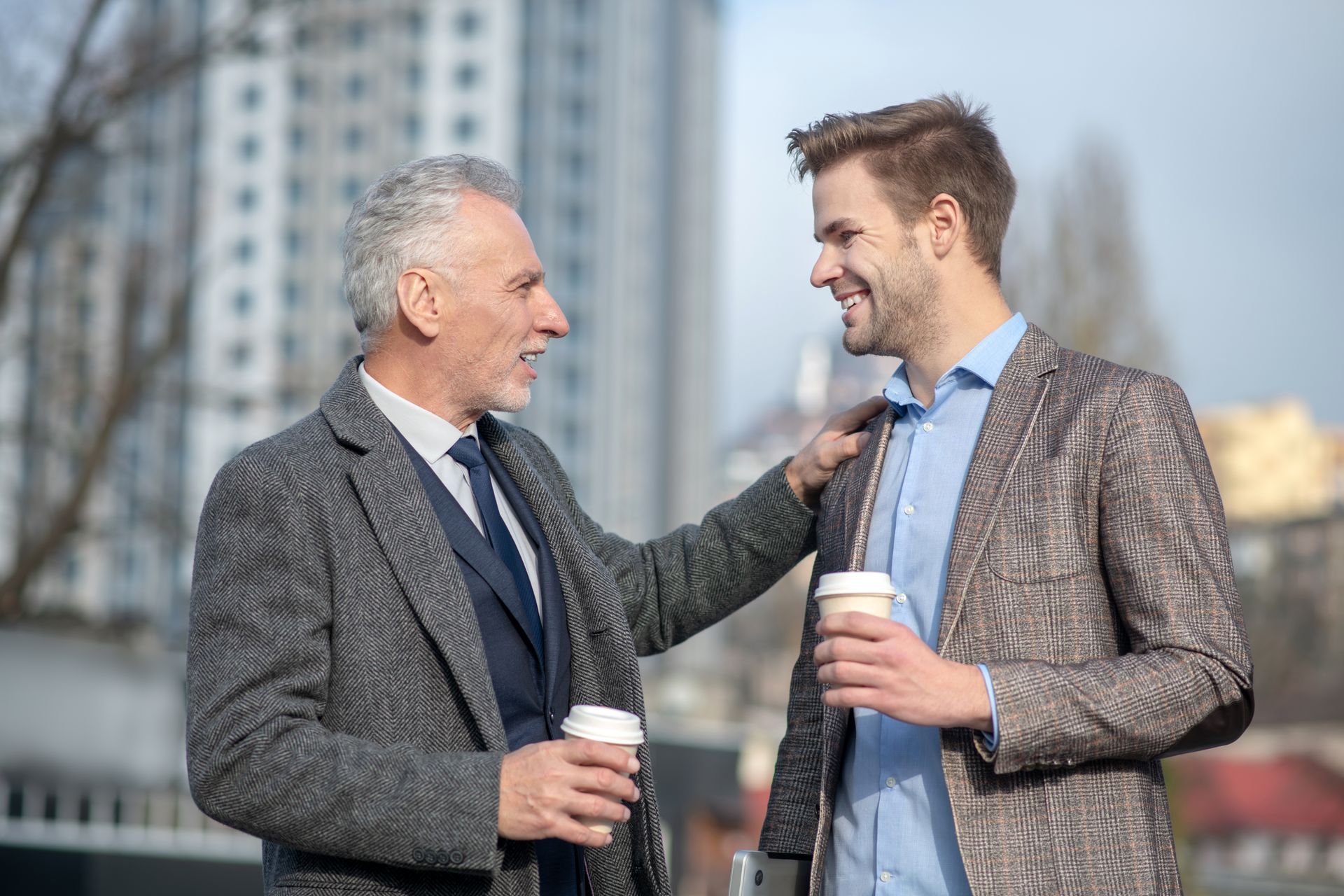 Two men chatting outdoors, smiling and holding coffee cups in a city setting.