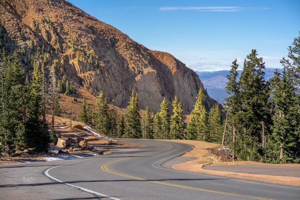 Winding mountain road through pine forest beside rocky cliffs under a clear blue sky