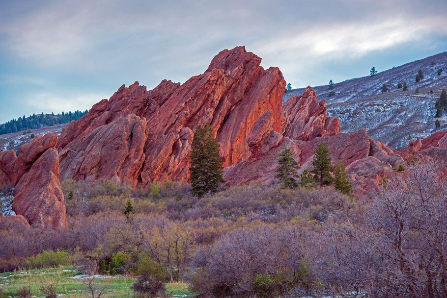 Red sandstone cliffs above purple shrubs and a green meadow under a cloudy sky