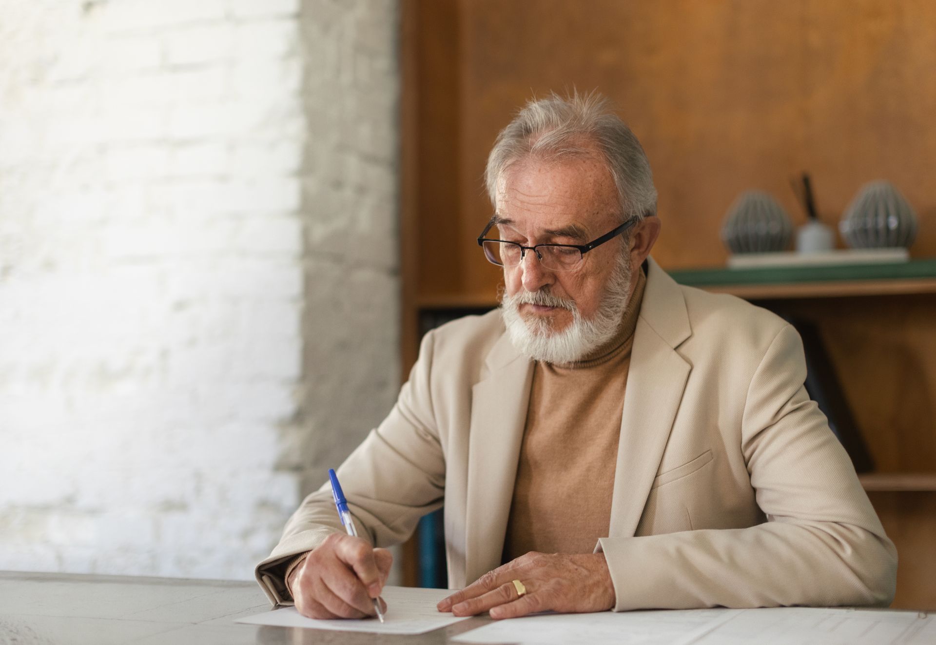 Man in a beige blazer writing at a desk beside a window