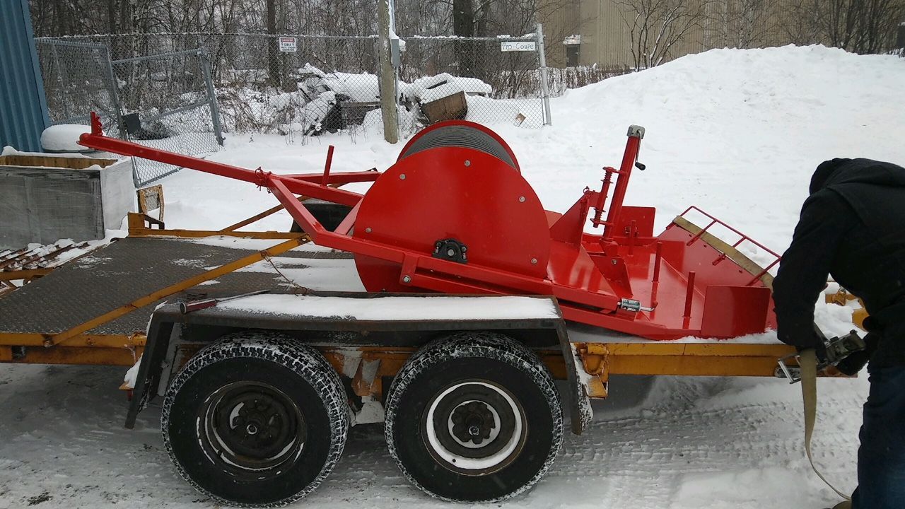 Souffleuse à neige rouge sur une remorque dans un décor enneigé. Une personne la fixe avec une sangle.
