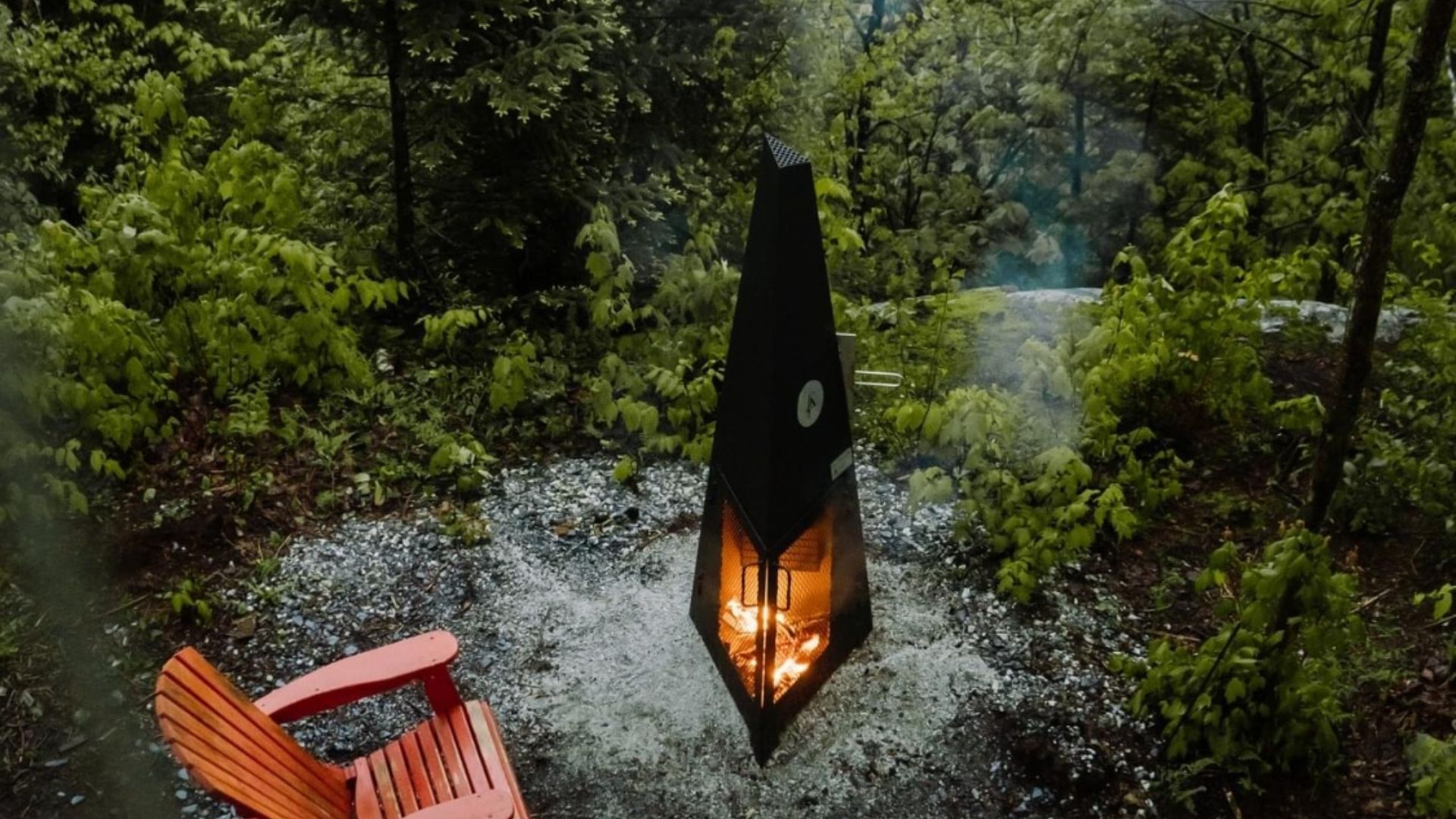 Cheminée extérieure noire allumée avec une chaise orange sous la pluie ; entourée de feuillage vert.