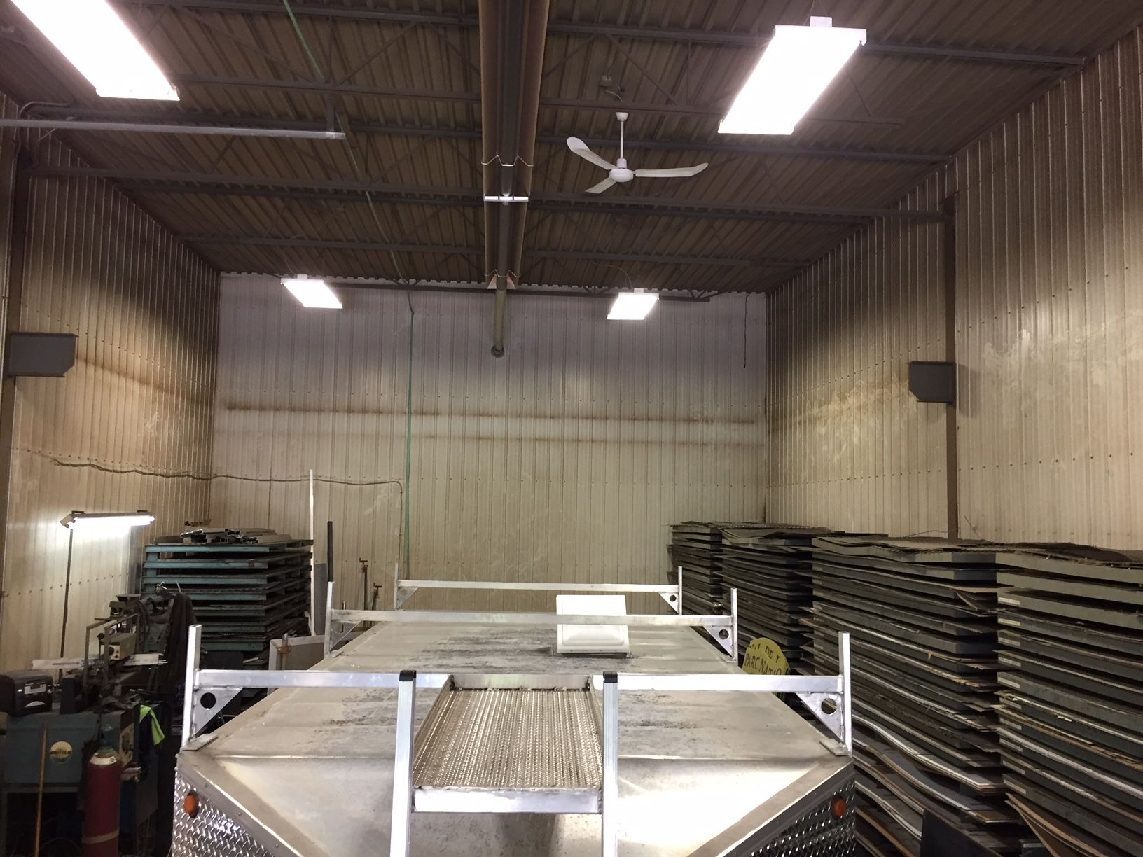 Un atelier industriel vide avec une plate-forme de camion au premier plan, les murs sont en métal, un éclairage zénithal et un ventilateur de plafond.