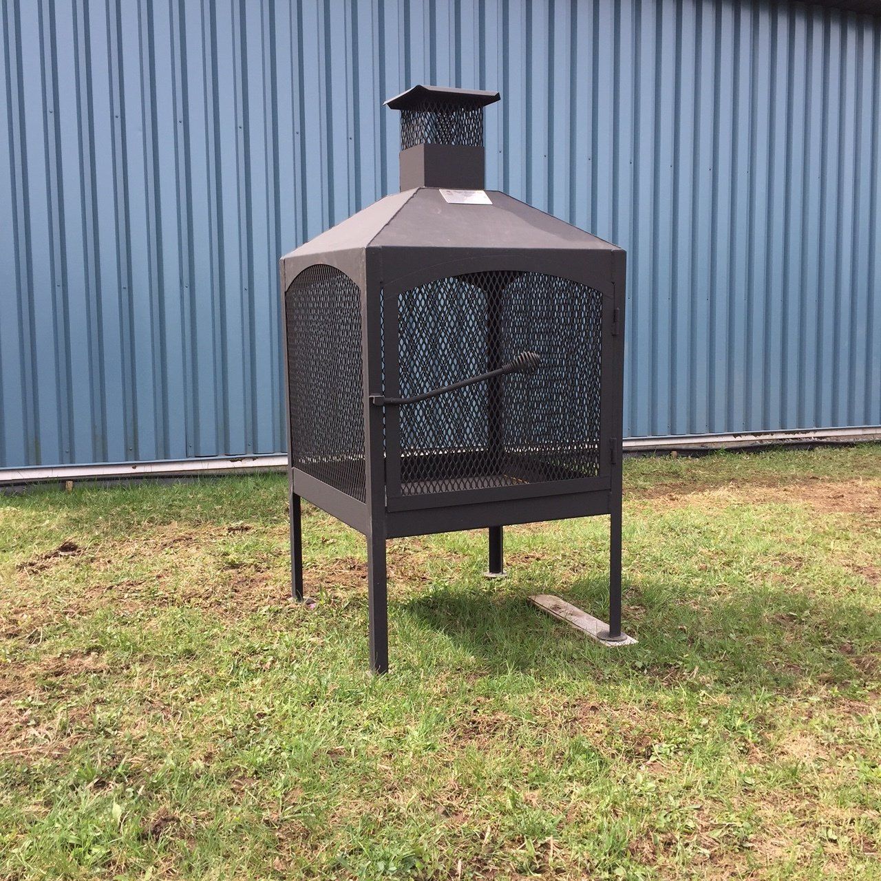 Foyer extérieur en métal noir avec une enceinte en treillis métallique, devant un bâtiment bleu.