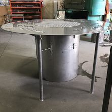 Table de barbecue d'extérieur en acier inoxydable avec plateau rond et base cylindrique, dans un cadre d'atelier.