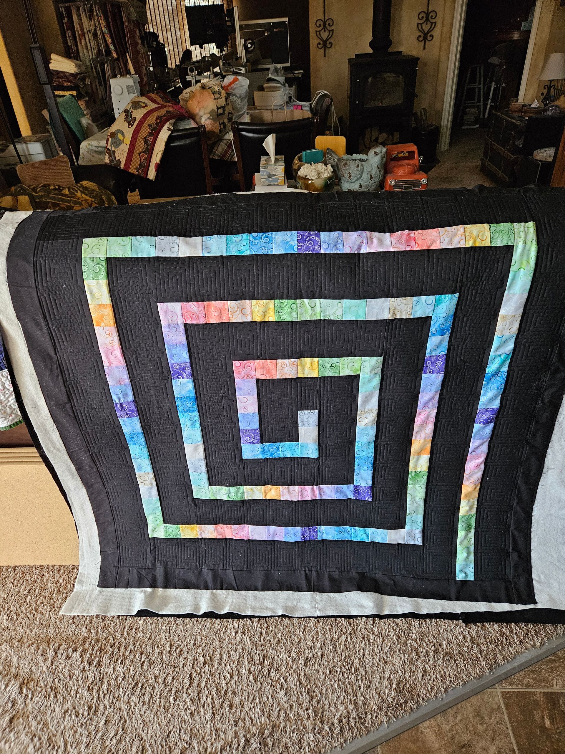 Black quilt with concentric spiral square pattern in rainbow colors, hanging on a chair indoors