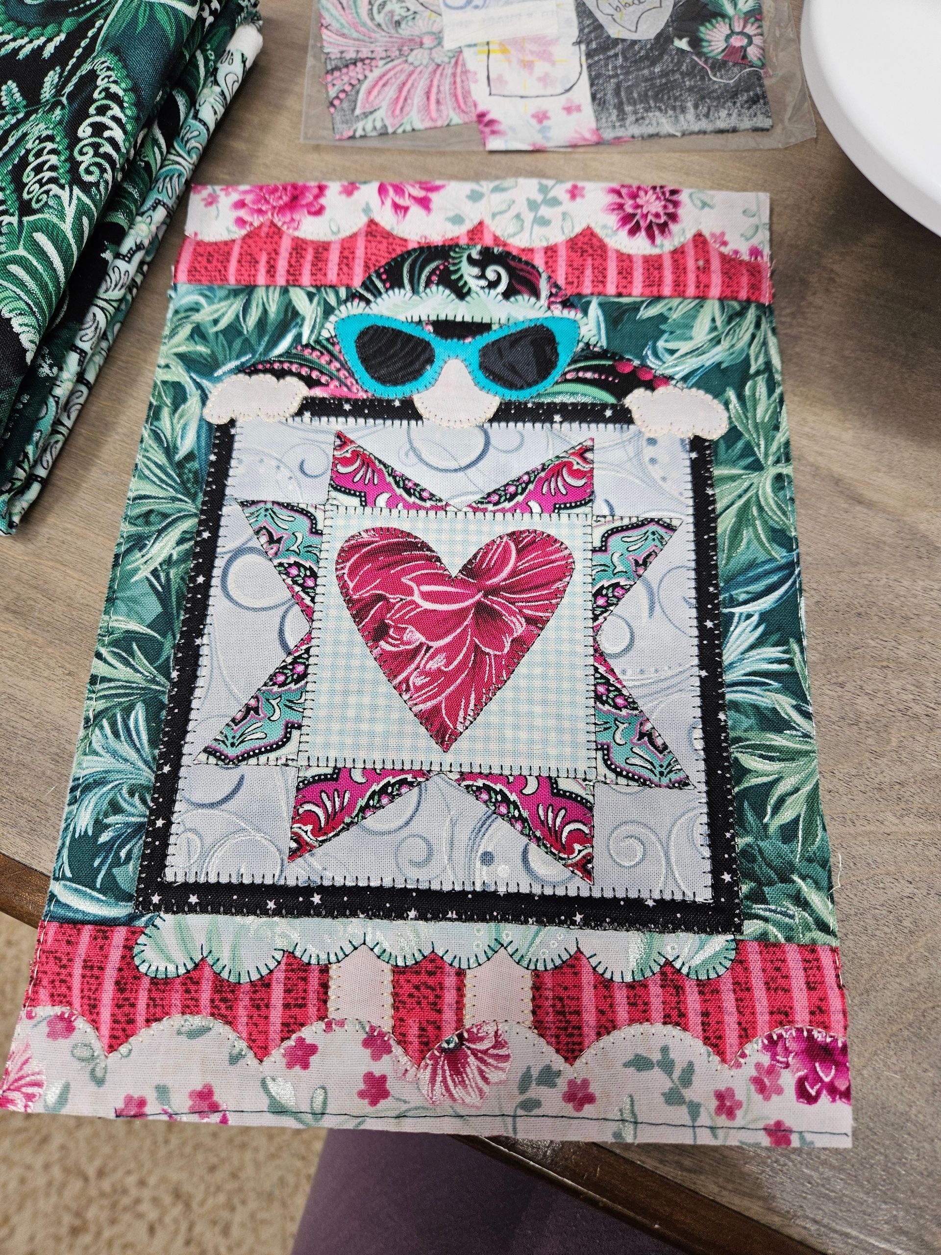 Patchwork quilt with a red heart and blue sunglasses on a bed or table, surrounded by floral fabric.