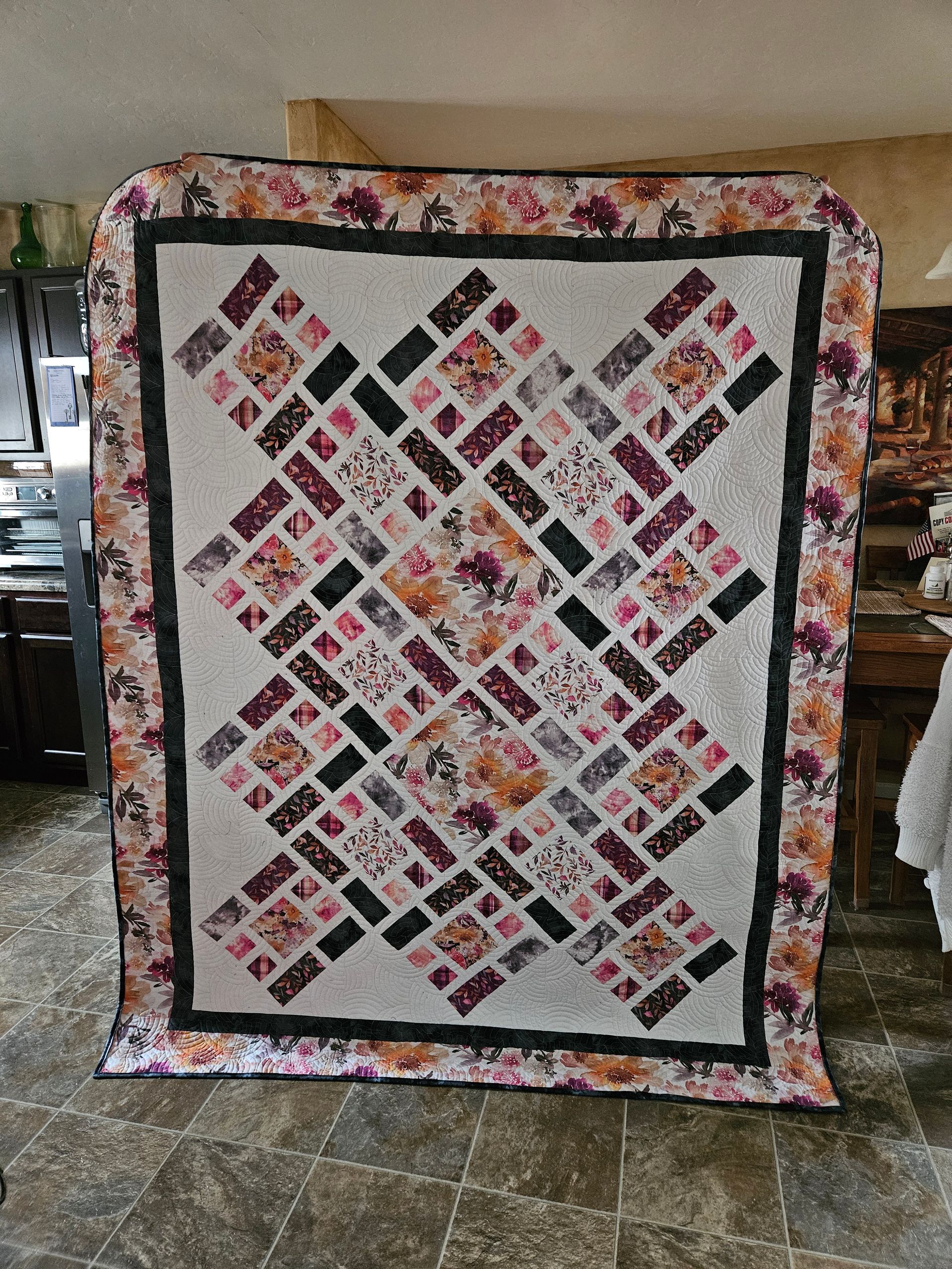 Floral patchwork quilt with diamond-patterned squares and dark border hanging indoors