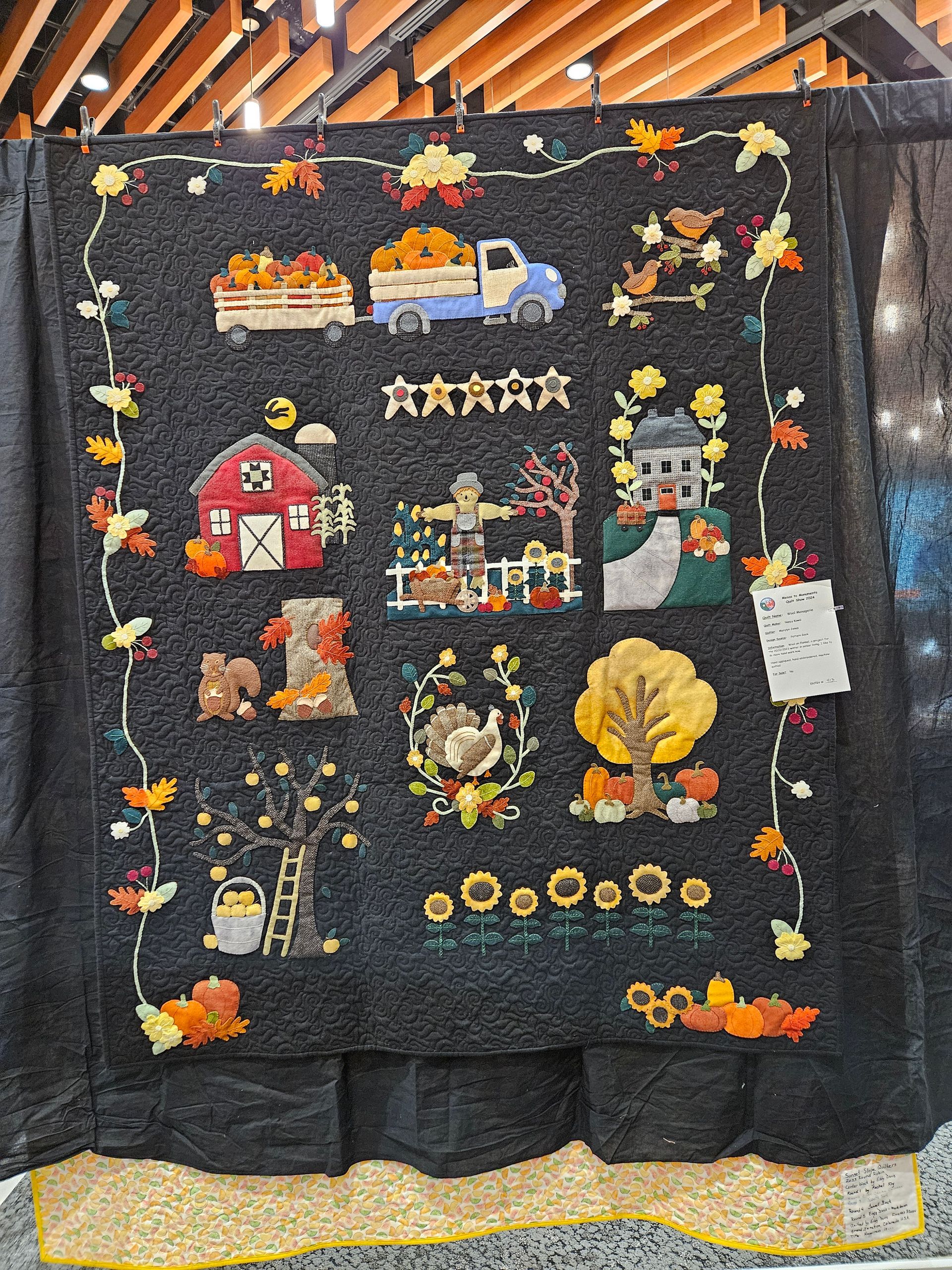 Black quilt with colorful embroidered festive scenes, stars, and hearts hanging on a wooden rack
