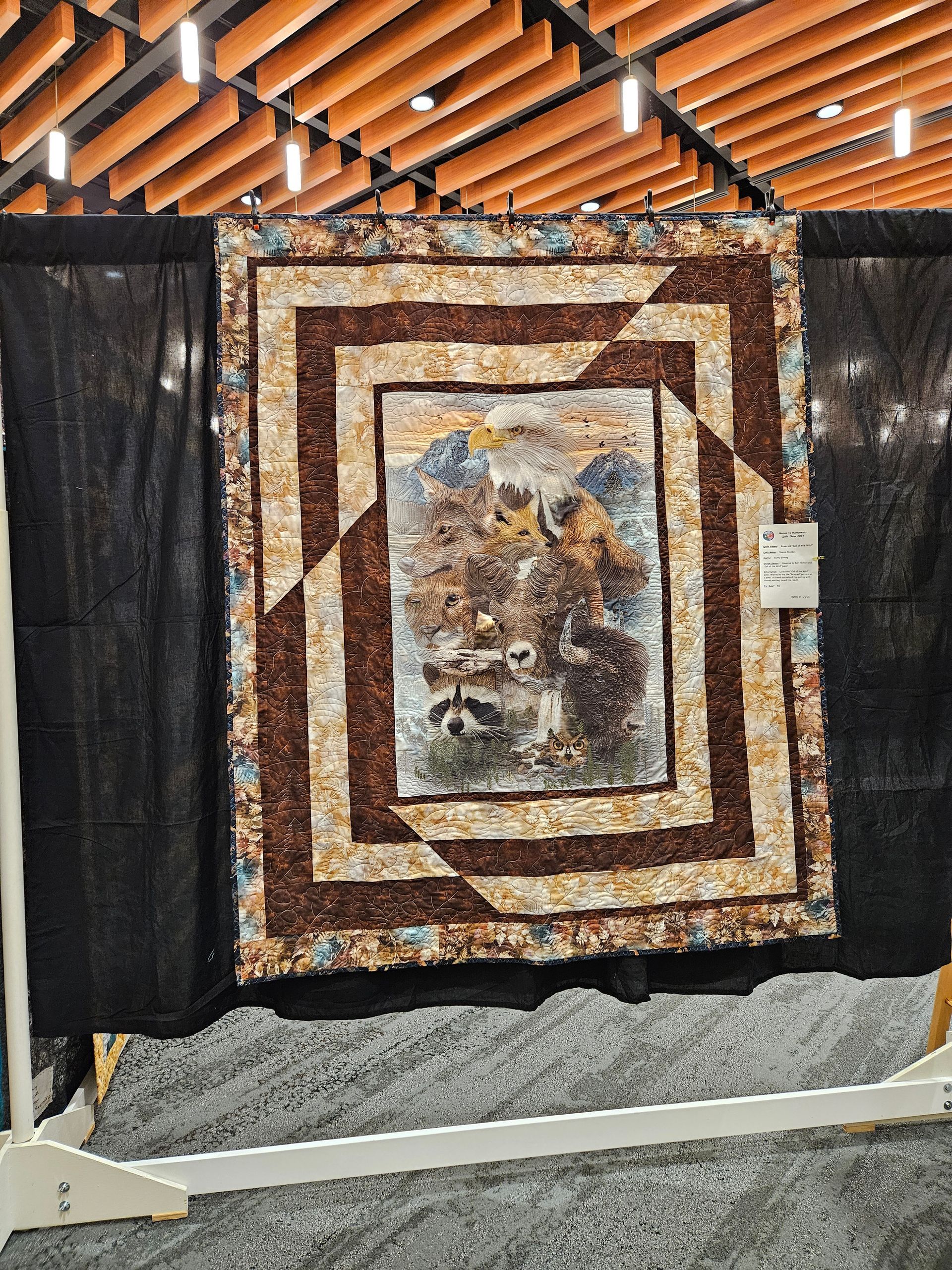 Quilt display with a tan and brown geometric quilt hanging indoors beneath a wooden roof.