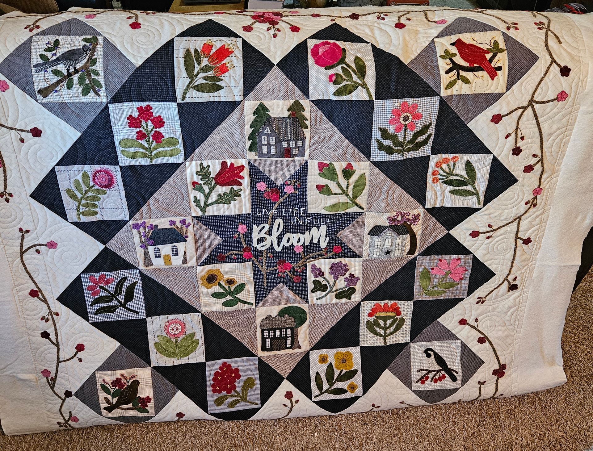 Quilt with a central “Bloom” panel, floral square patches, and vine border in pink, red, green, and black.