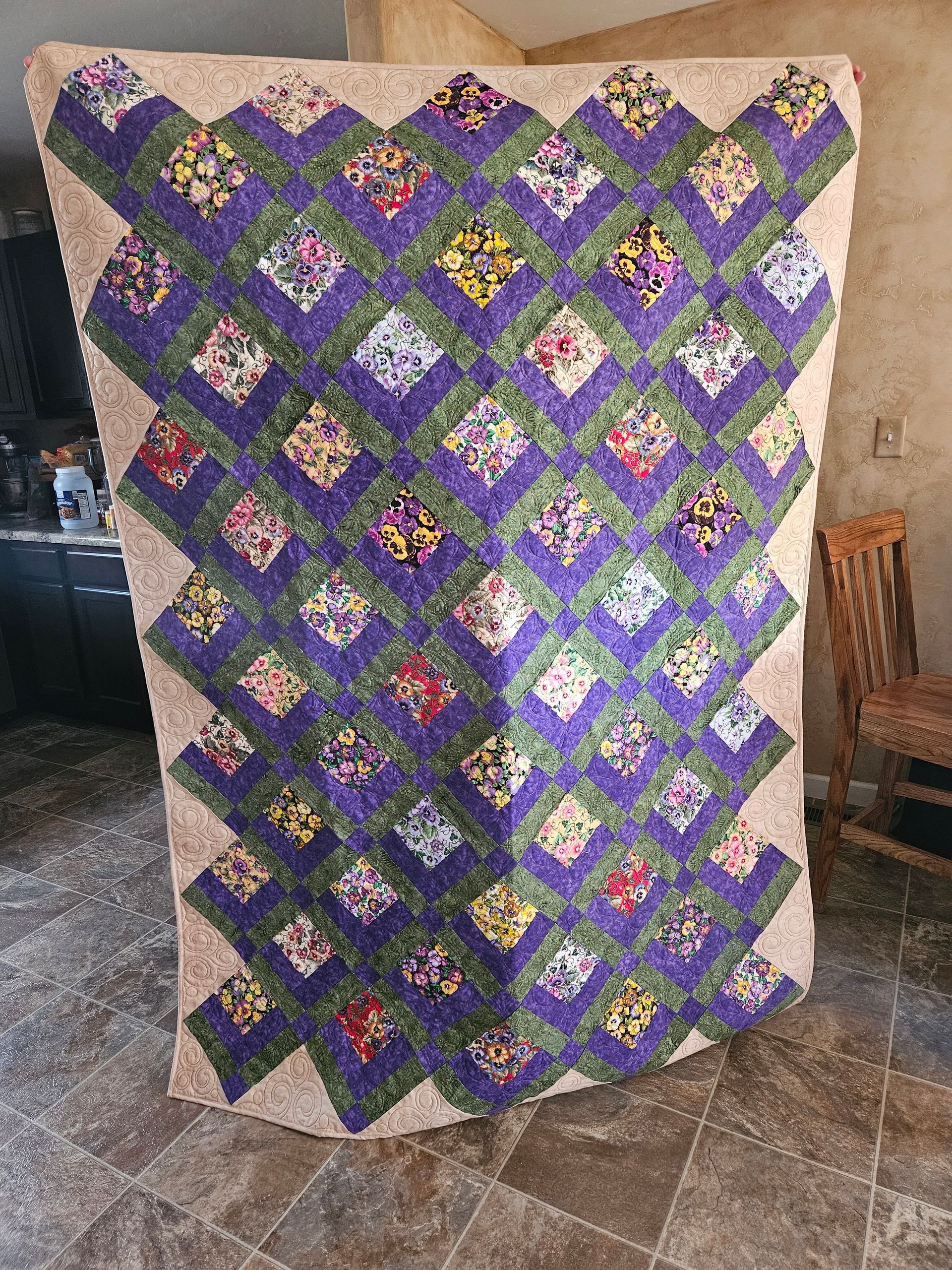 Quilt with purple, green, and beige diamond patchwork draped over a chair in a room.