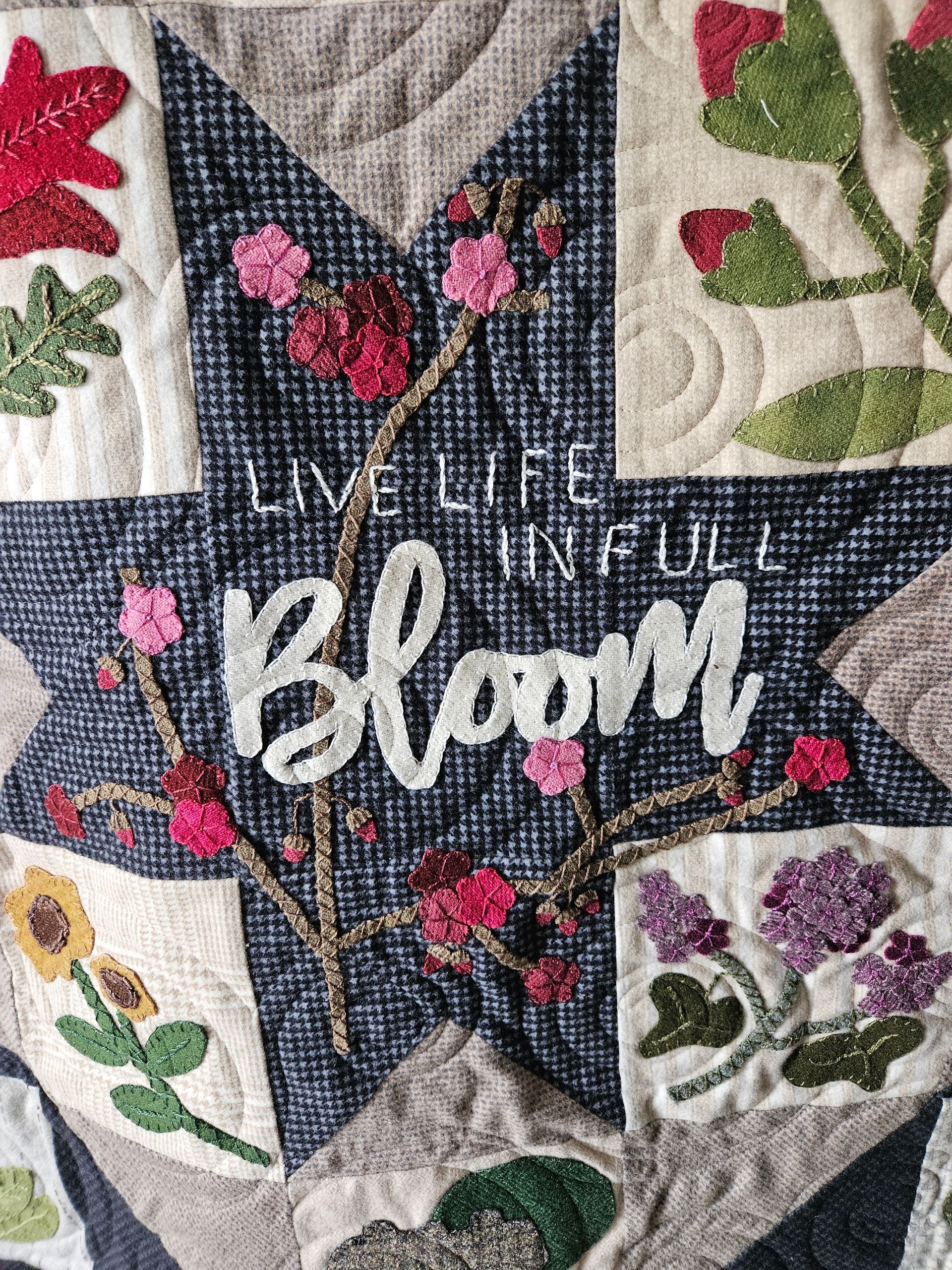 Quilt patch with dark blue star and white “Bloom” text, surrounded by floral embroidery in pink, green, and purple