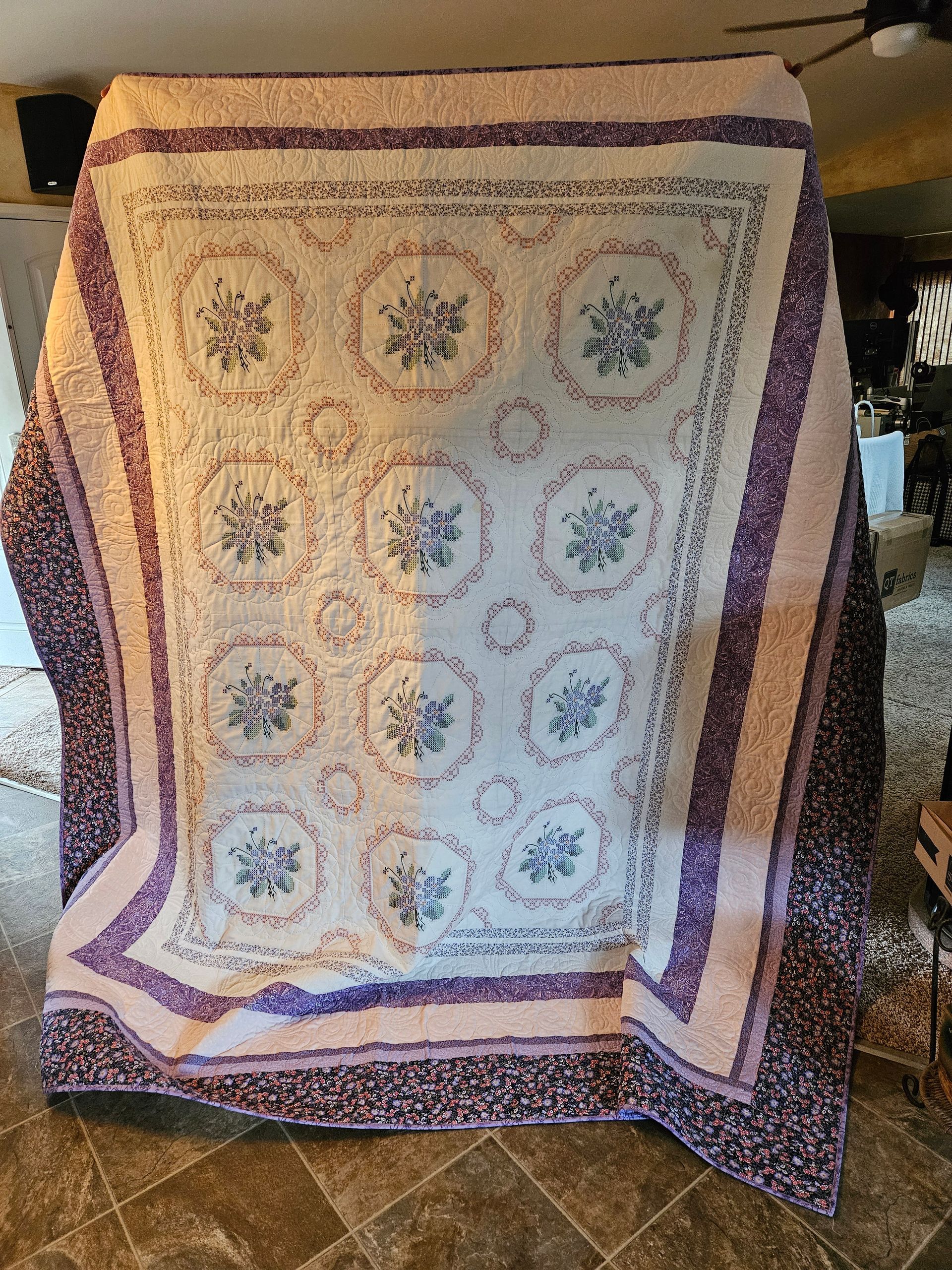 Patchwork quilt with pastel circular medallions, bordered in purple and draped over a stand indoors