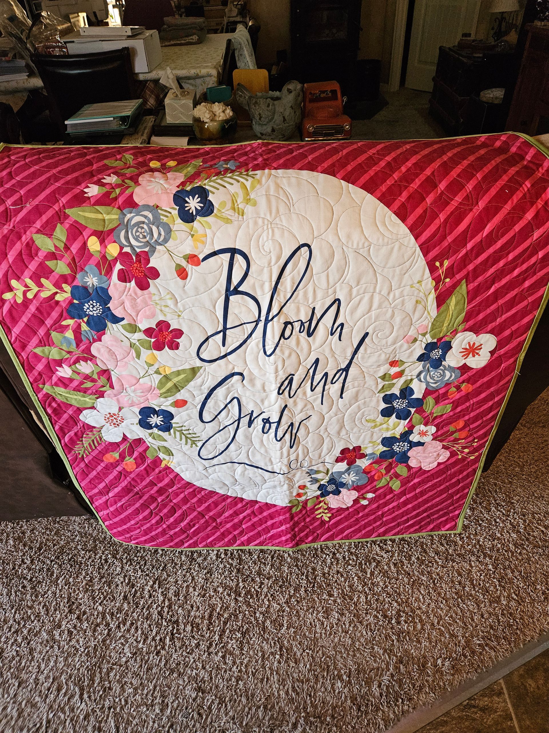 Floral “Bless this Grand” sign with pink, white, and red diamond-patterned border on a couch.