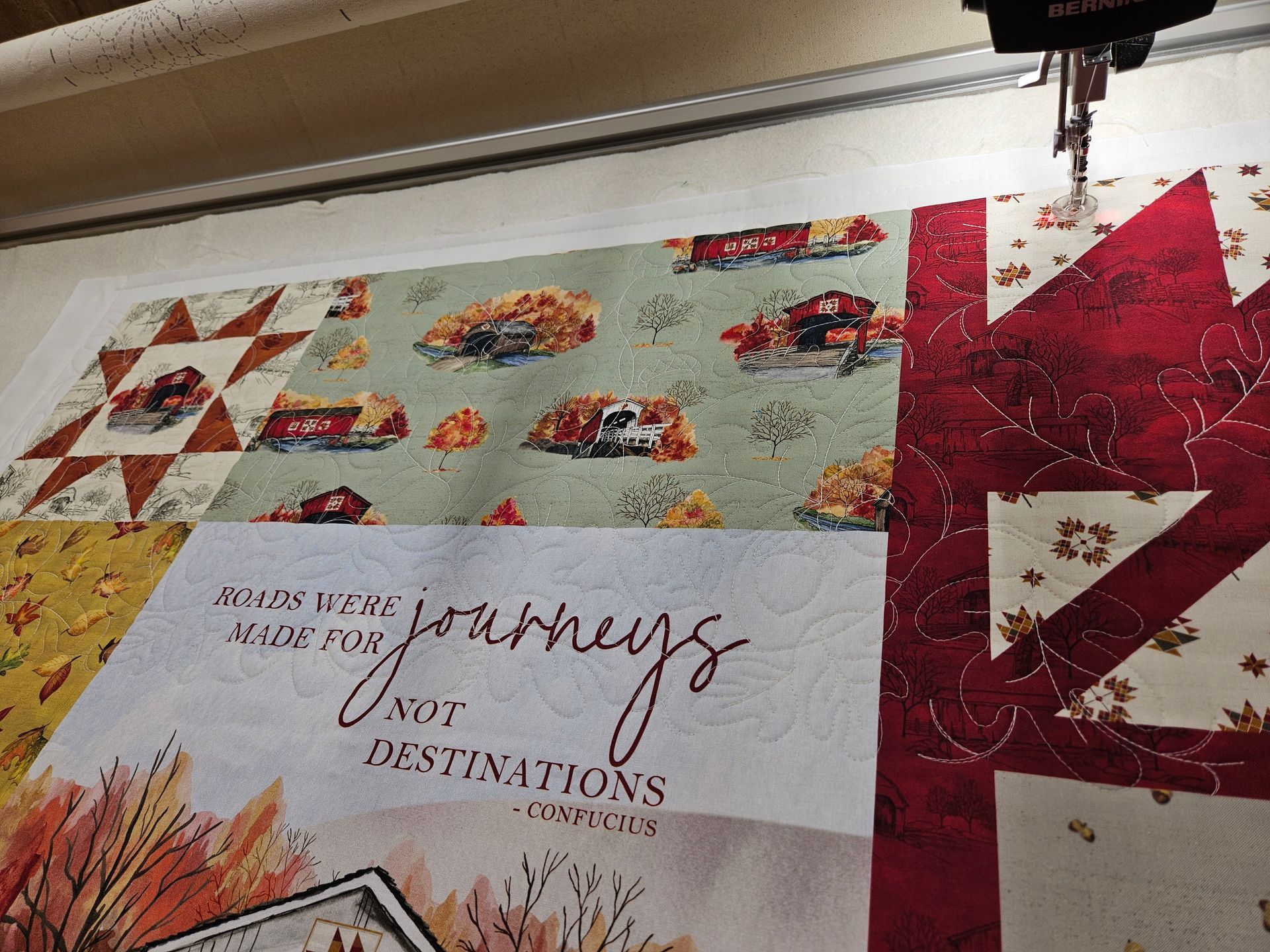 Quilt top with red, cream, and floral blocks, featuring a central “journeys” panel under a sewing machine