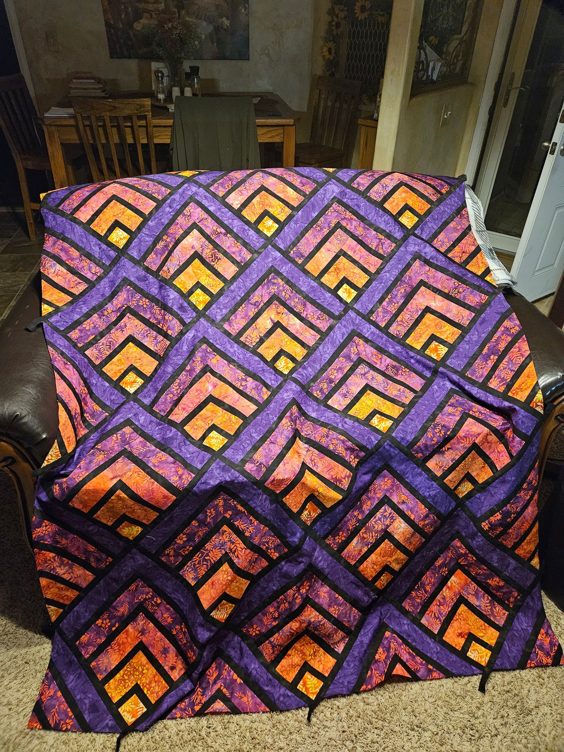 Colorful purple, orange, and black geometric quilt draped over a chair in a dim room