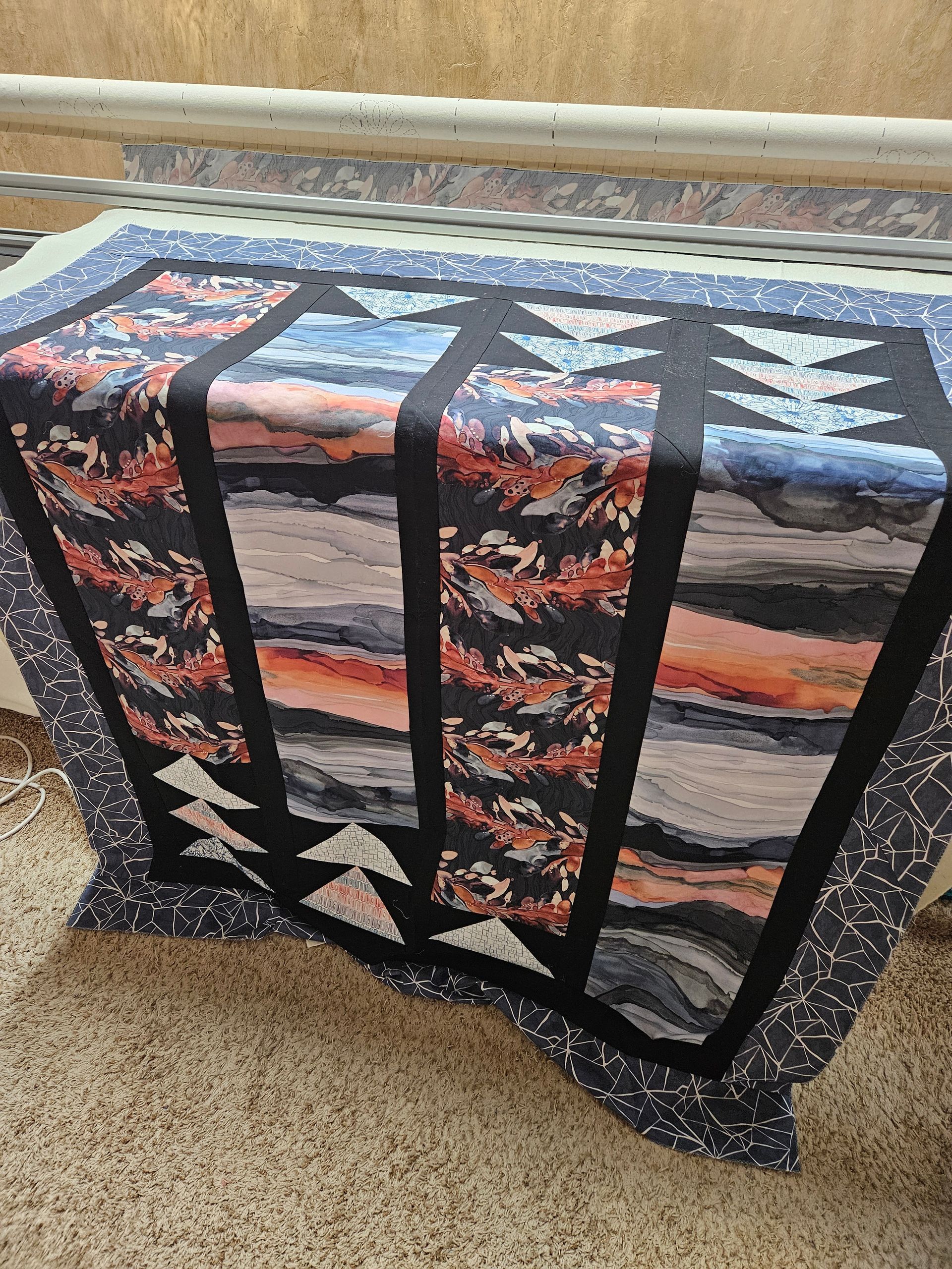 Quilted table runner with black border and sunset-and-tree patchwork draped over a beige carpet.