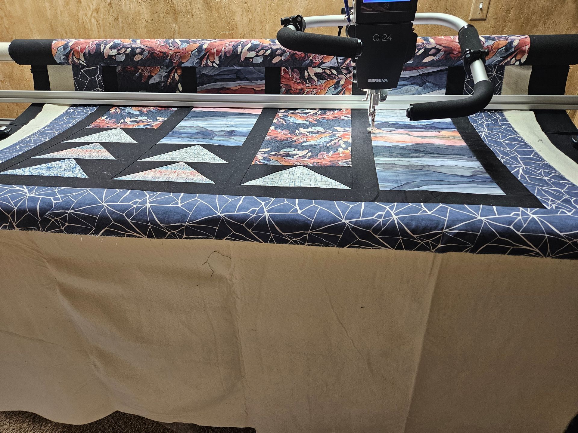 Quilt top under a longarm quilting machine, with geometric fabric patterns and a tan table cover.
