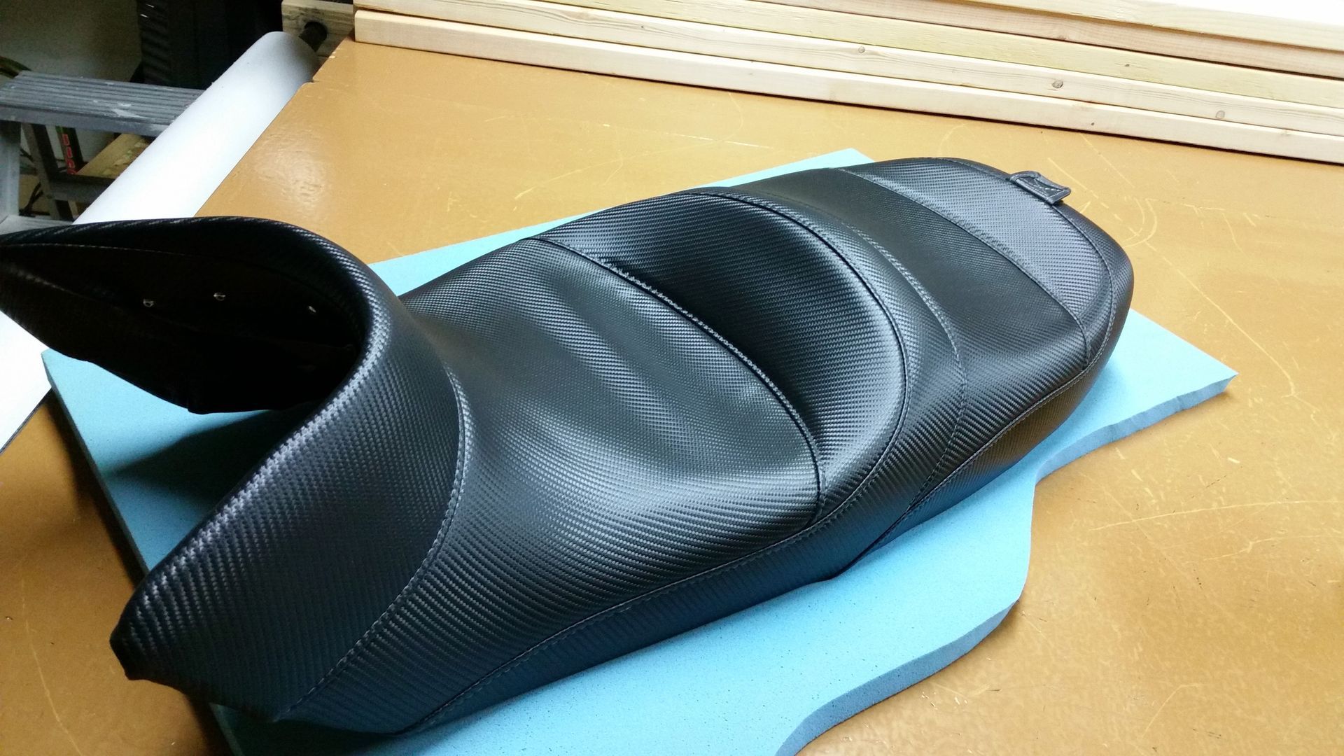 Black carbon-fiber motorcycle seat on a blue mat in a workshop-like setting