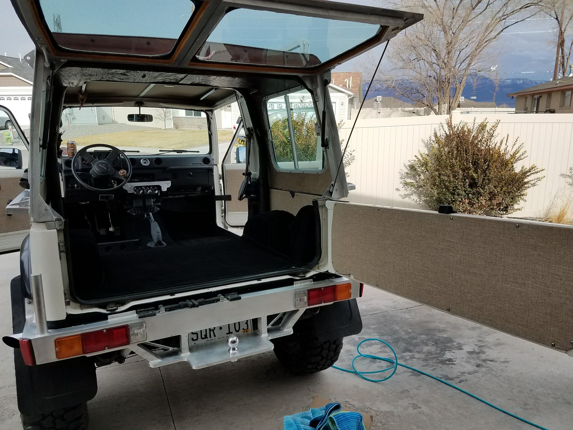 Open back of a white SUV with rear seats removed, parked on a driveway near a fence and mountains.