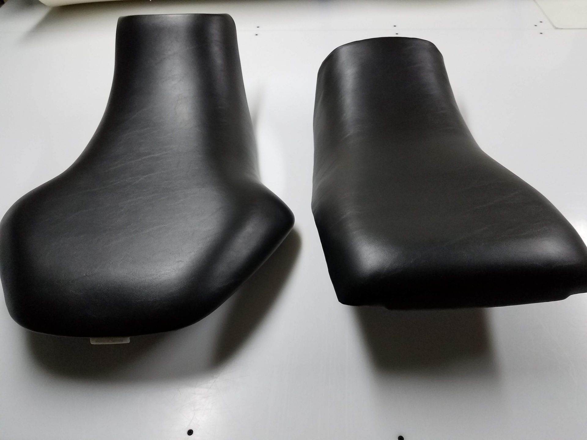 Two black motorcycle seats on a white surface, shown side by side.