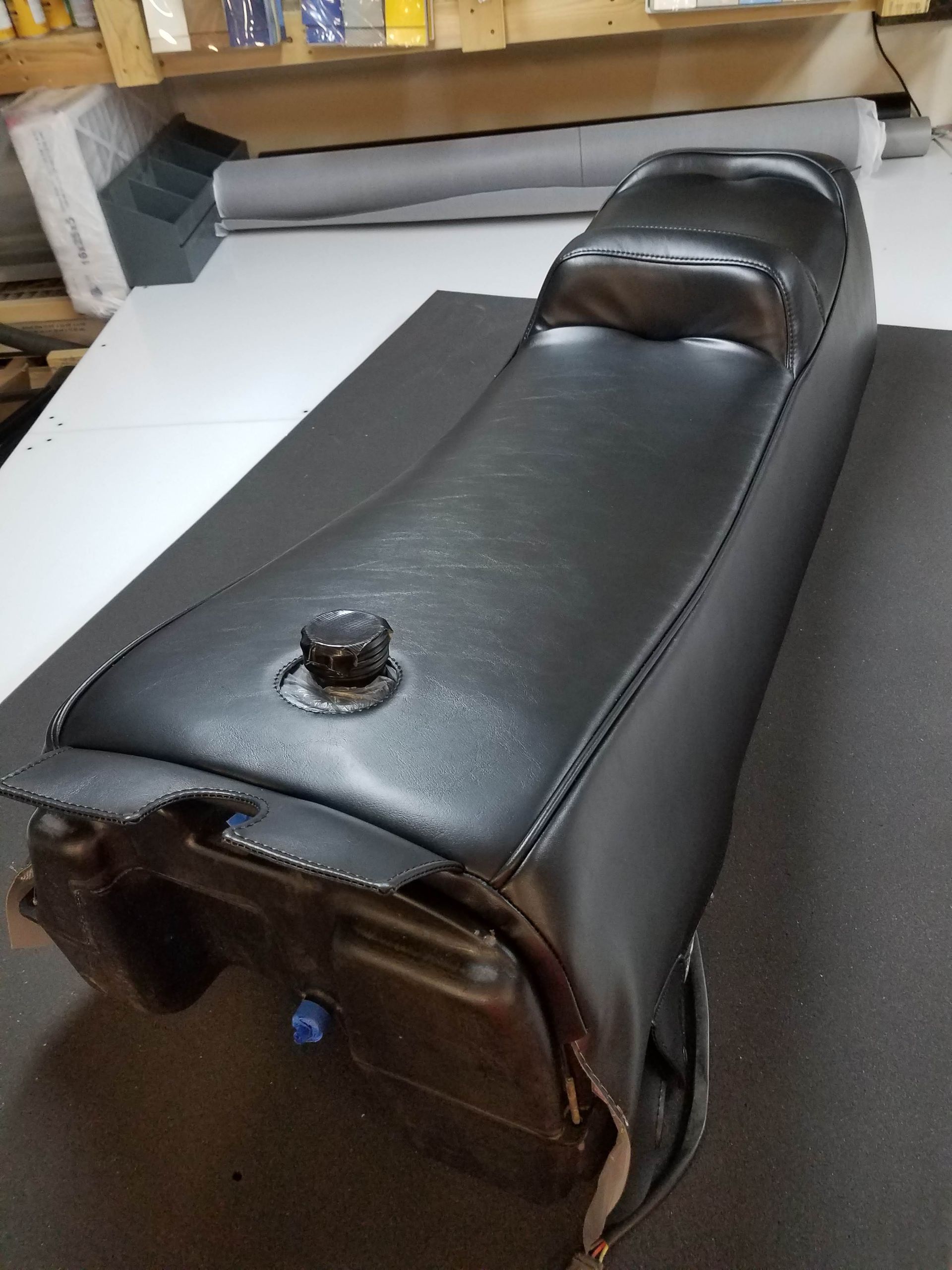 Dark brown leather motorcycle seat on a workshop table, with a fuel cap visible and a gray roll in back.