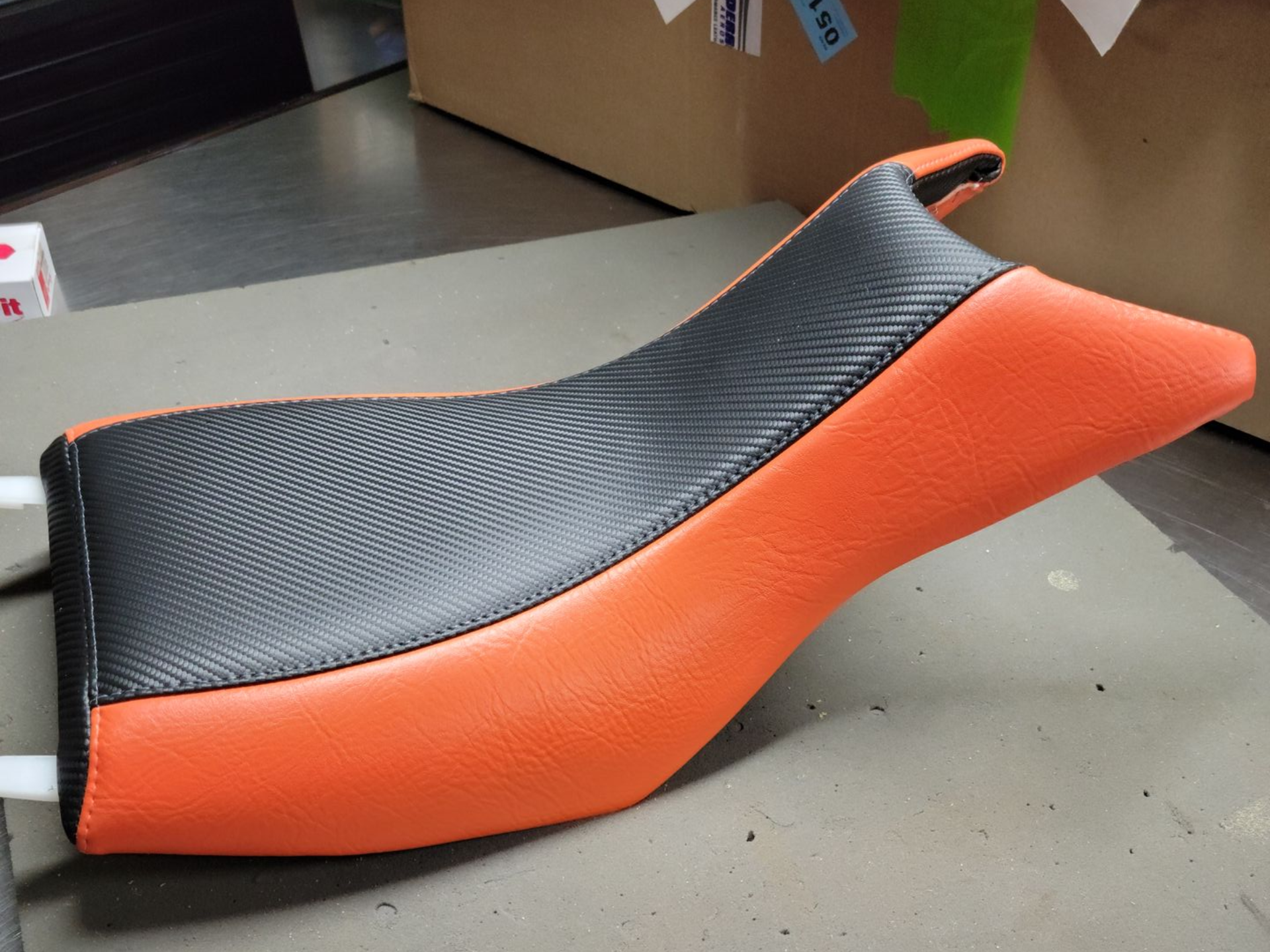 Black and orange motorcycle seat with a textured black top, shown on a gray floor