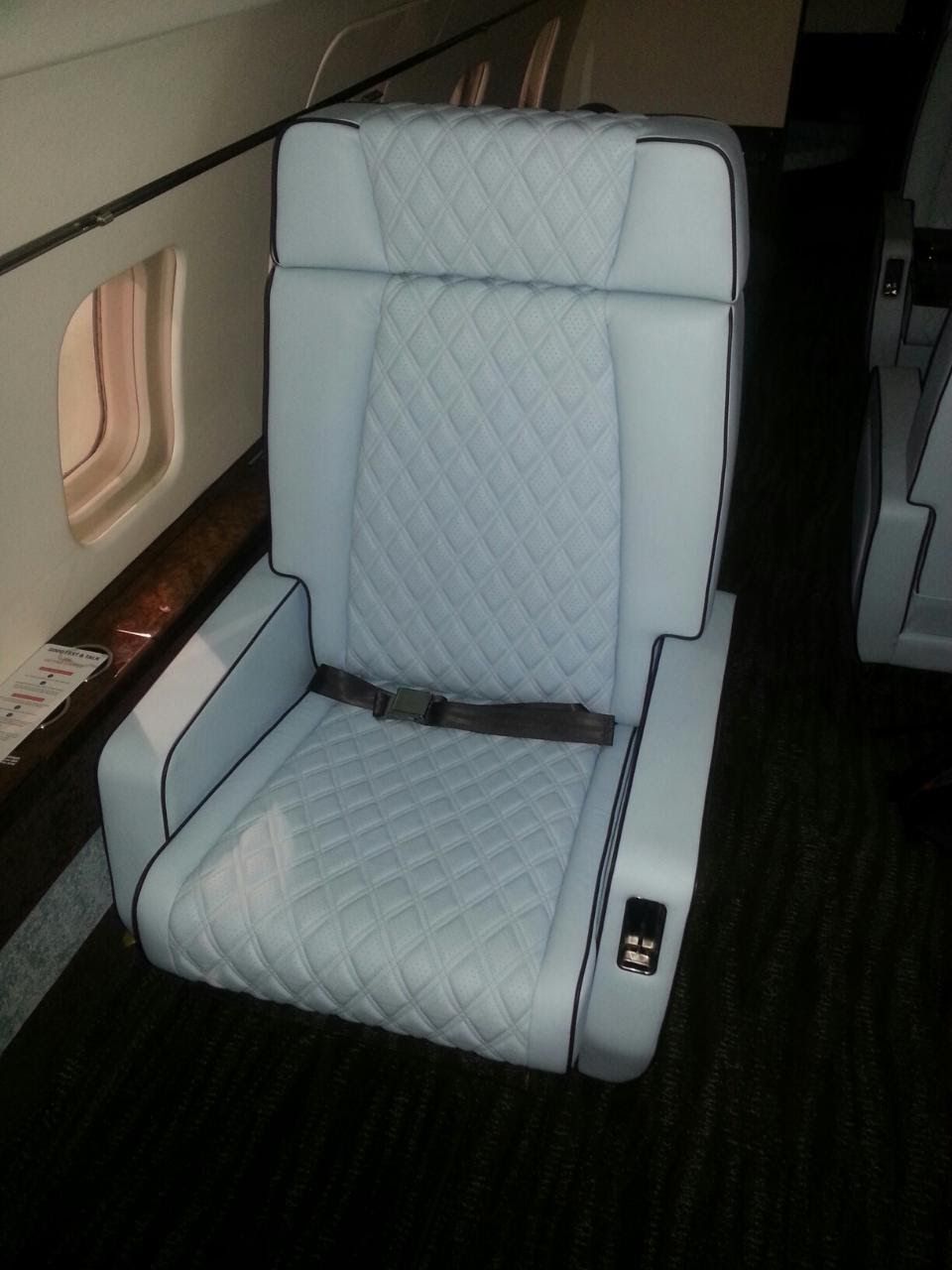 Light blue upholstered airplane seat with armrests and seatbelt, beside a small window.