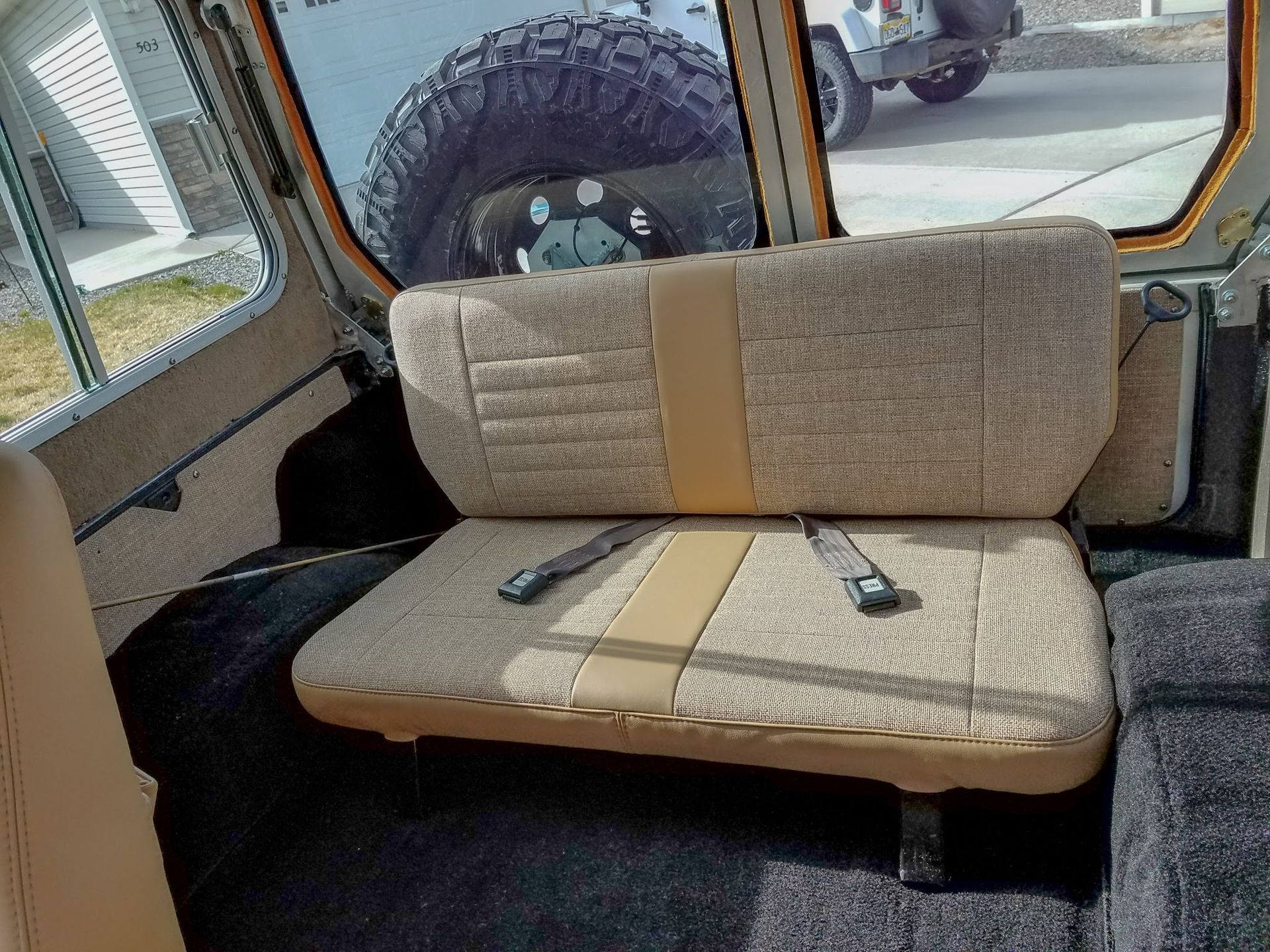 Beige bench seat inside a vehicle cab, with seat belts and a rear window visible.