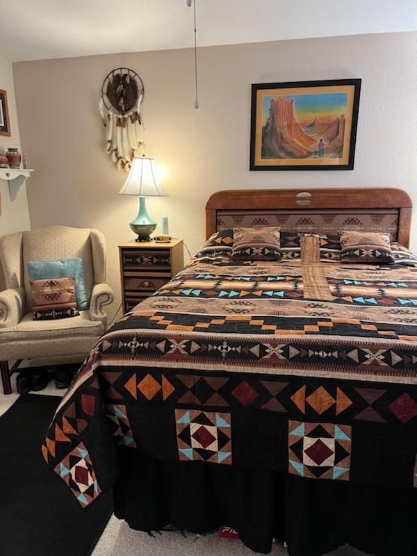 Cozy bedroom with a patterned quilt, wooden bed, armchair, lamp, and dreamcatcher on the wall.