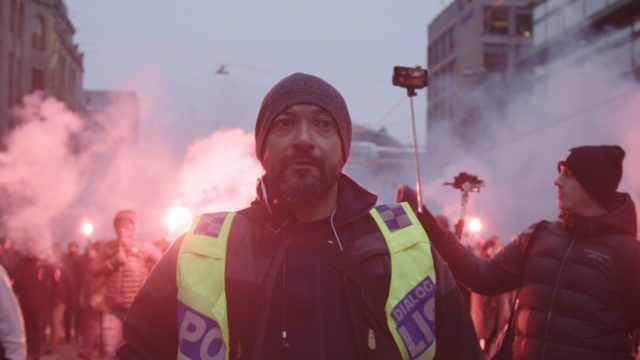 A man in a yellow vest is standing in front of a crowd of people.