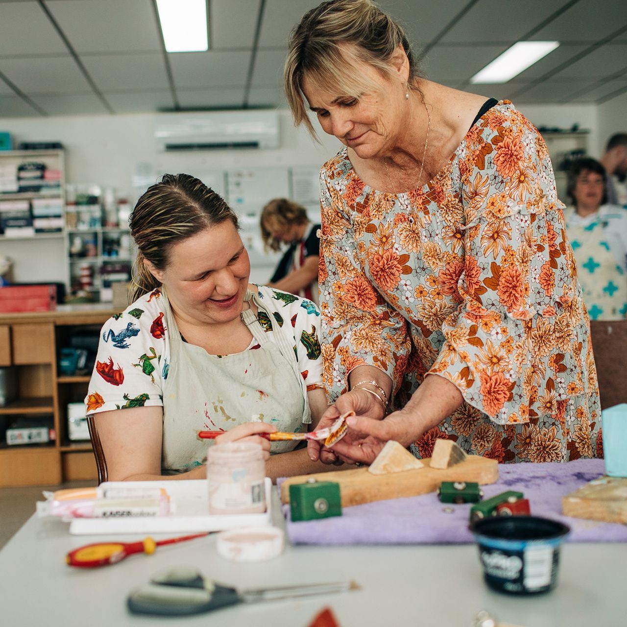 Woman assisting another painting wooden craft project at a table.