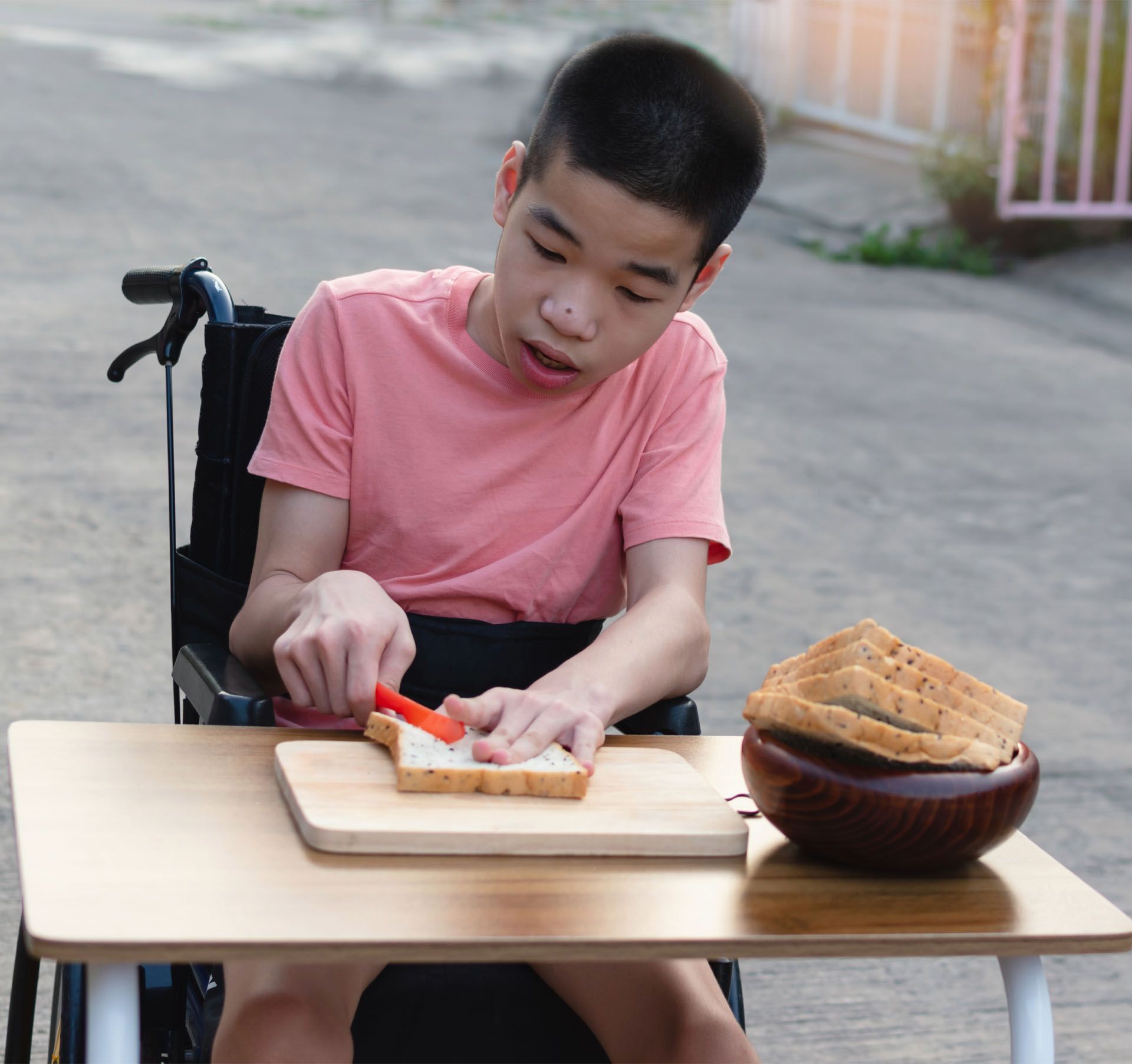 A young boy in a wheelchair is cutting bread on a cutting board