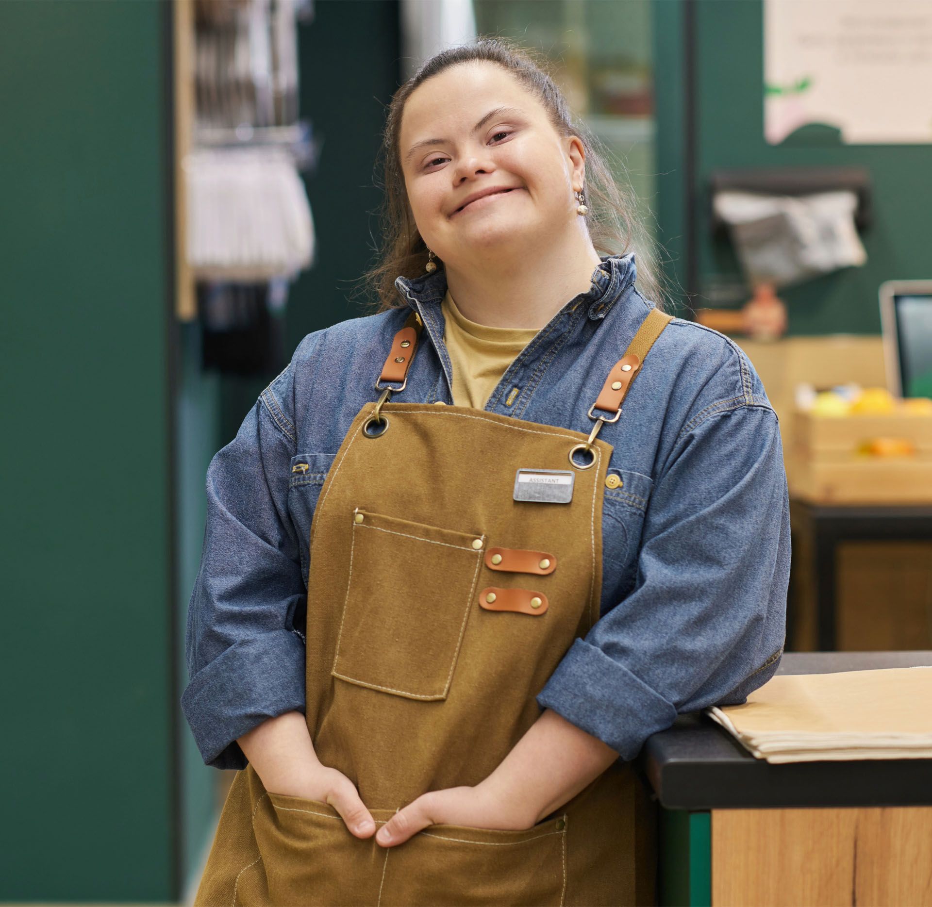 A woman wearing an apron and a denim shirt is standing in front of a counter.
