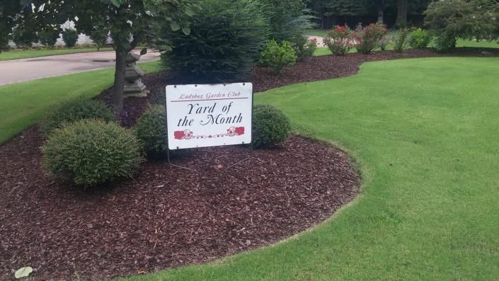 Lawn sign in a landscaped yard reading “In Loving Memory of Grandma,” with red flowers.