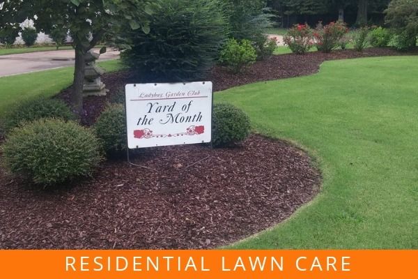 Lawn care yard sign in a landscaped front yard with mulch, shrubs, and green grass