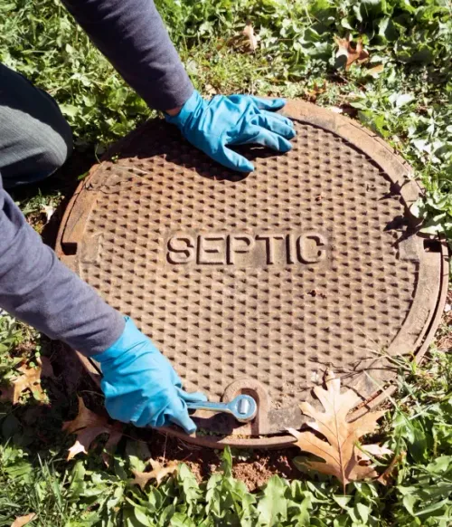 Gloved hands opening a round septic tank cover outdoors among grass and leaves