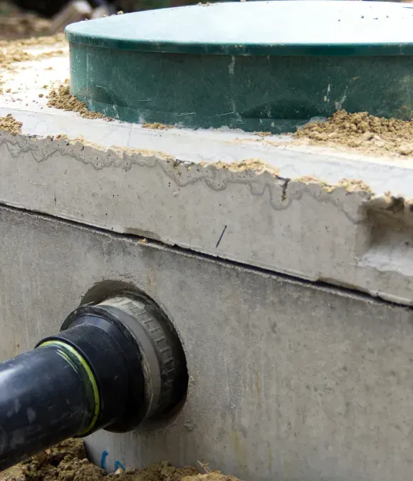 Black pipe entering a concrete tank wall below a green circular opening with debris around the rim