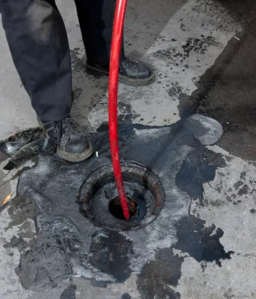 A red hose drains or clears a dark clogged hole in wet pavement beside workers’ boots.