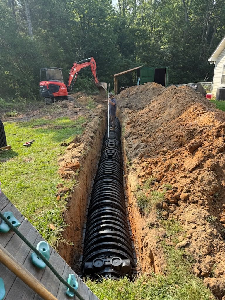Excavator installing black drainage pipe in a long trench beside a house and dirt piles