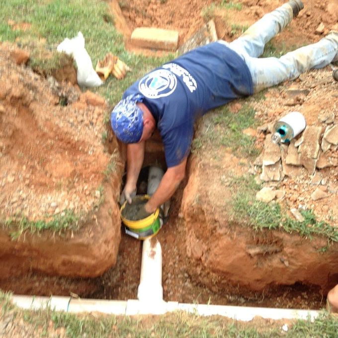 Worker in blue shirt using a tool in a narrow trench beside exposed white pipes.