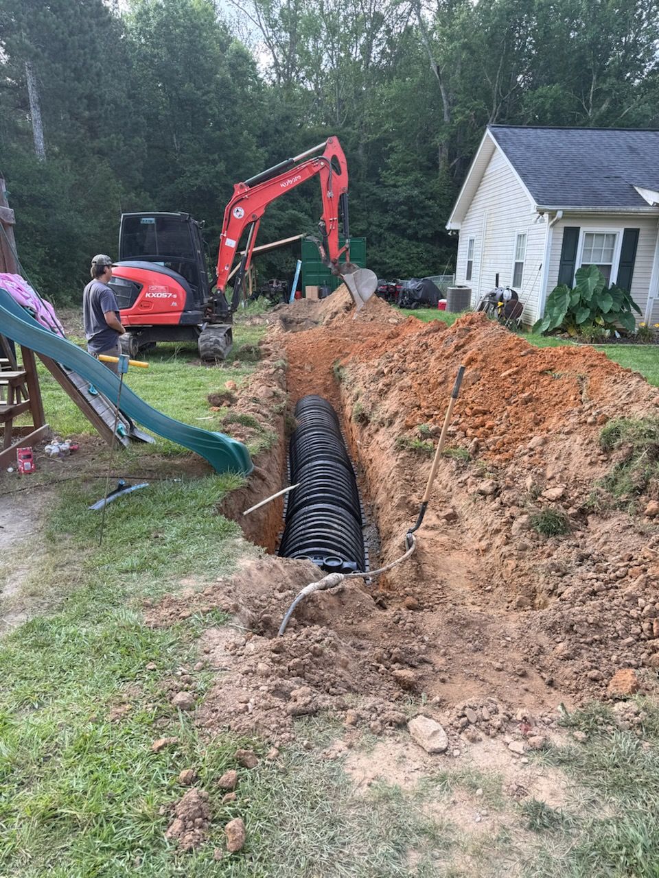 Excavator digging a trench beside a house, exposing black drainage pipe in a muddy yard.