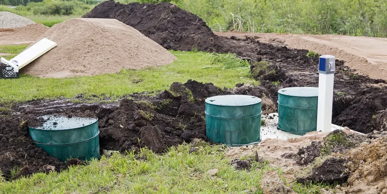 Utility tanks and a white pipe installed in a dirt excavation beside soil piles and grass.