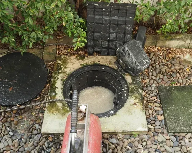 Open manhole with water, surrounded by gravel and a red pipe in a garden area