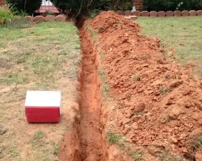A deep trench runs through a grassy yard beside a red-and-white cooler