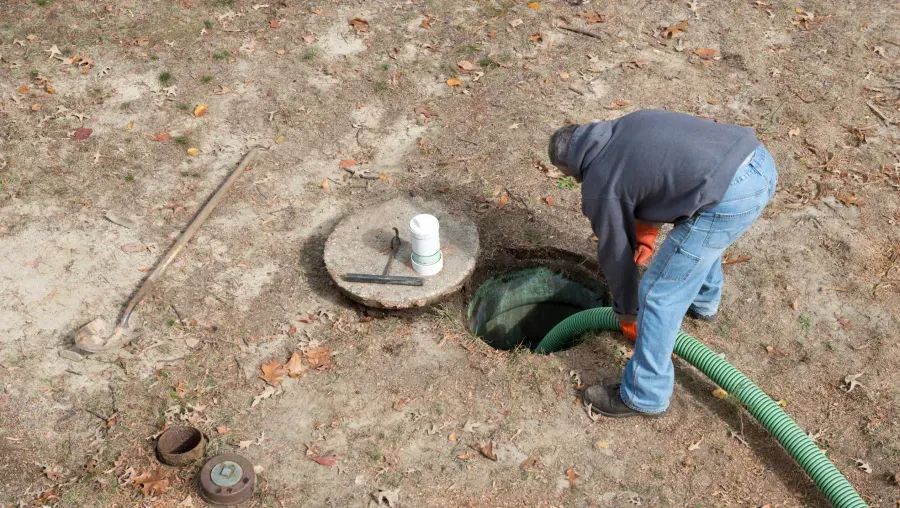 Worker bends over a green hose beside a circular utility cover in a dirt area outdoors.